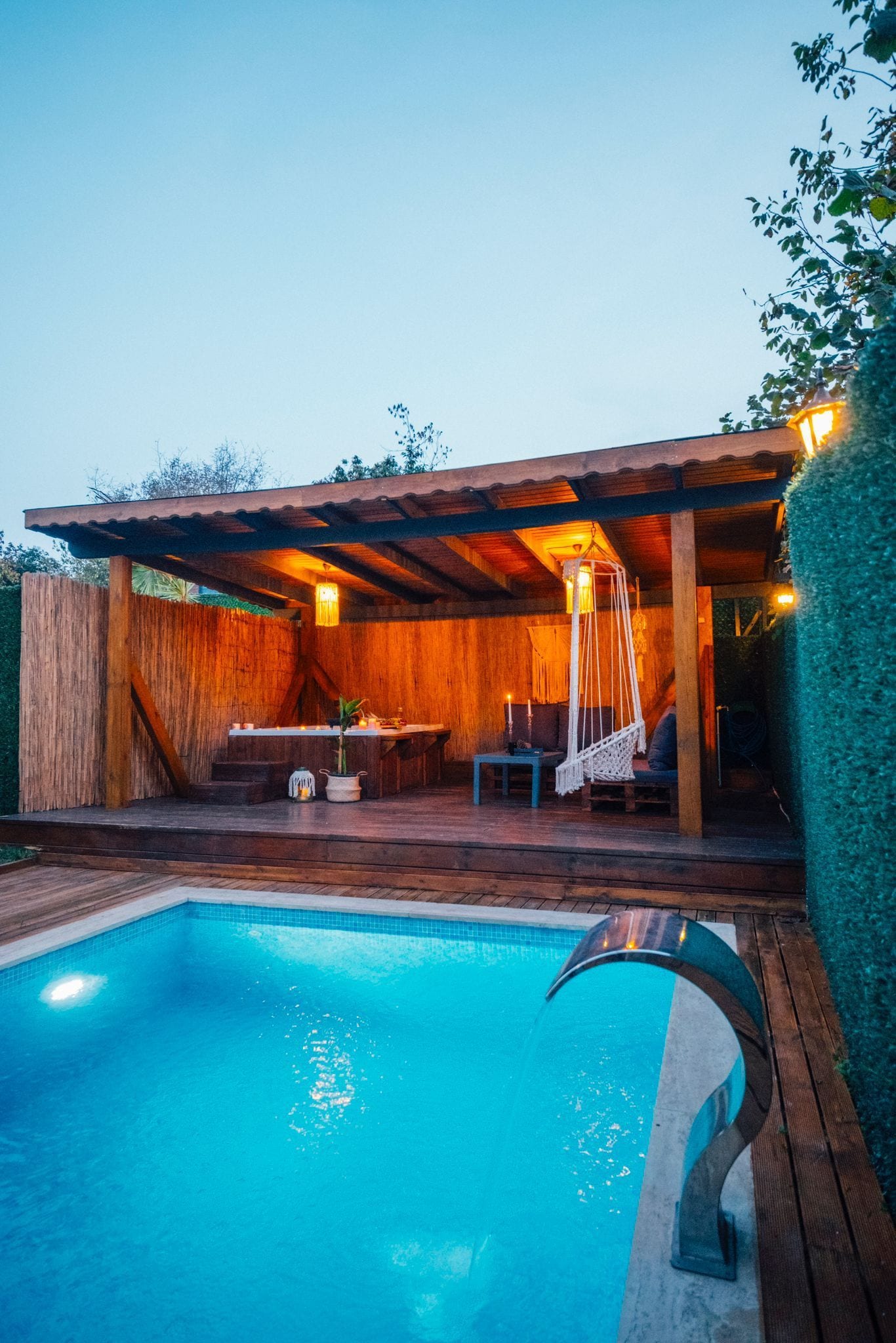 A swimming pool with a wooden deck, a covered seating area, and a hanging light fixture.