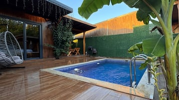 Ein Schwimmbecken mit Holzterrasse, einer Hängematte und einer überdachten Sitzgruppe.