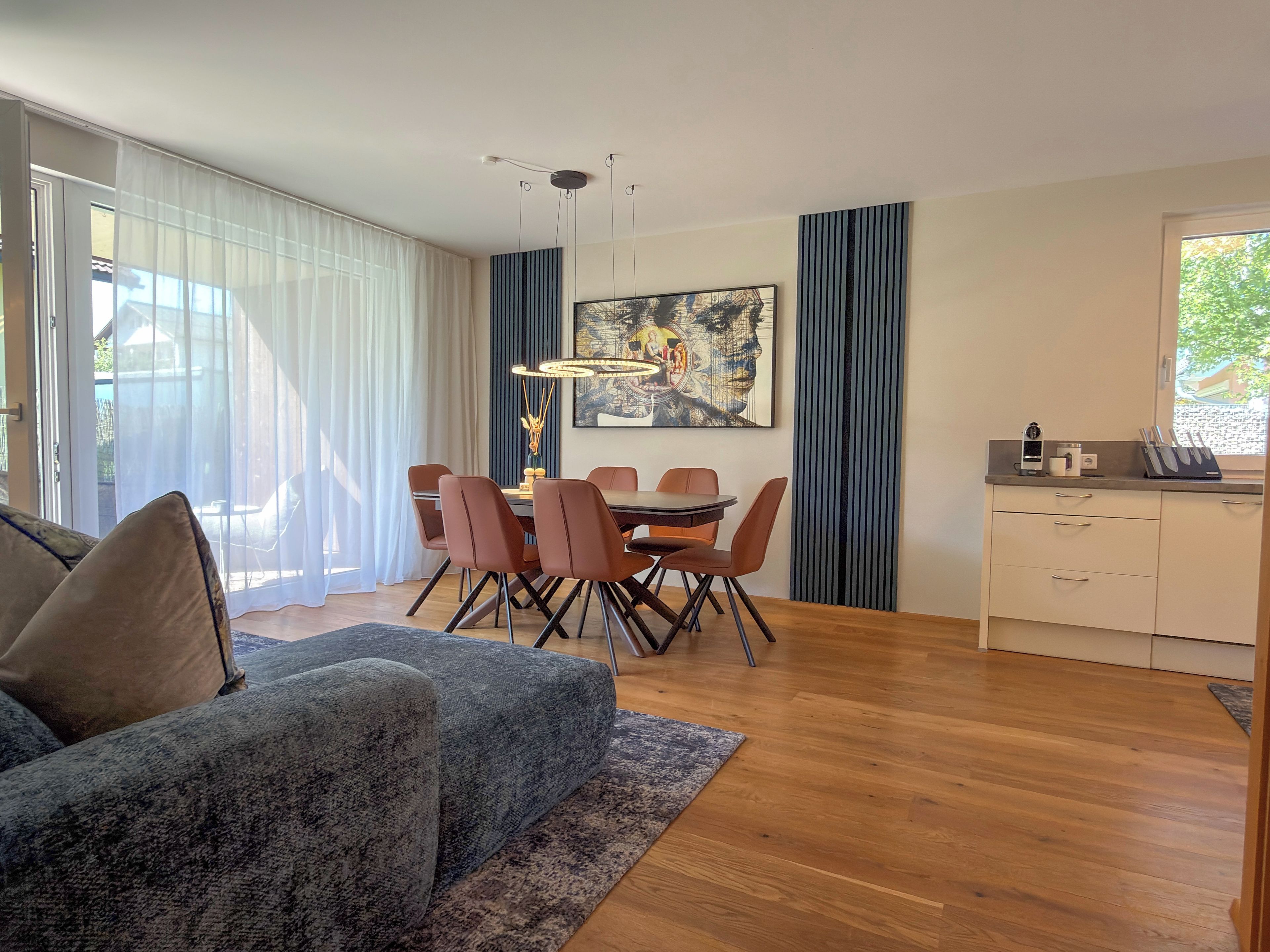 A modern living room with a sofa, a dining table with chairs, a kitchen area, and a large window with curtains.