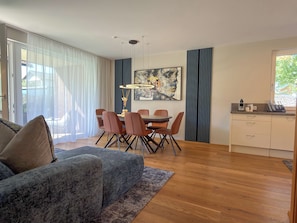A modern living room with a sofa, a dining table with chairs, a kitchen area, and a large window with curtains.