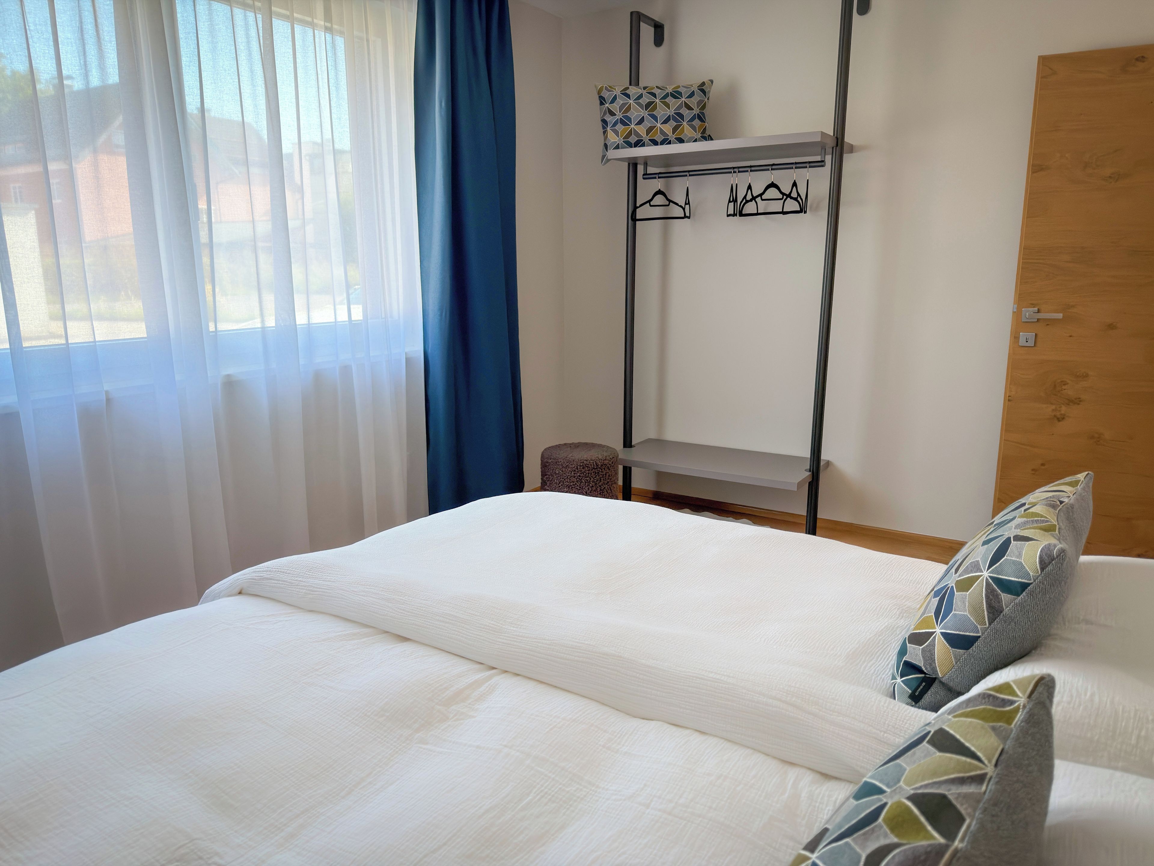 A bed with a white duvet, a metal shelf above it, and a patterned pillow.