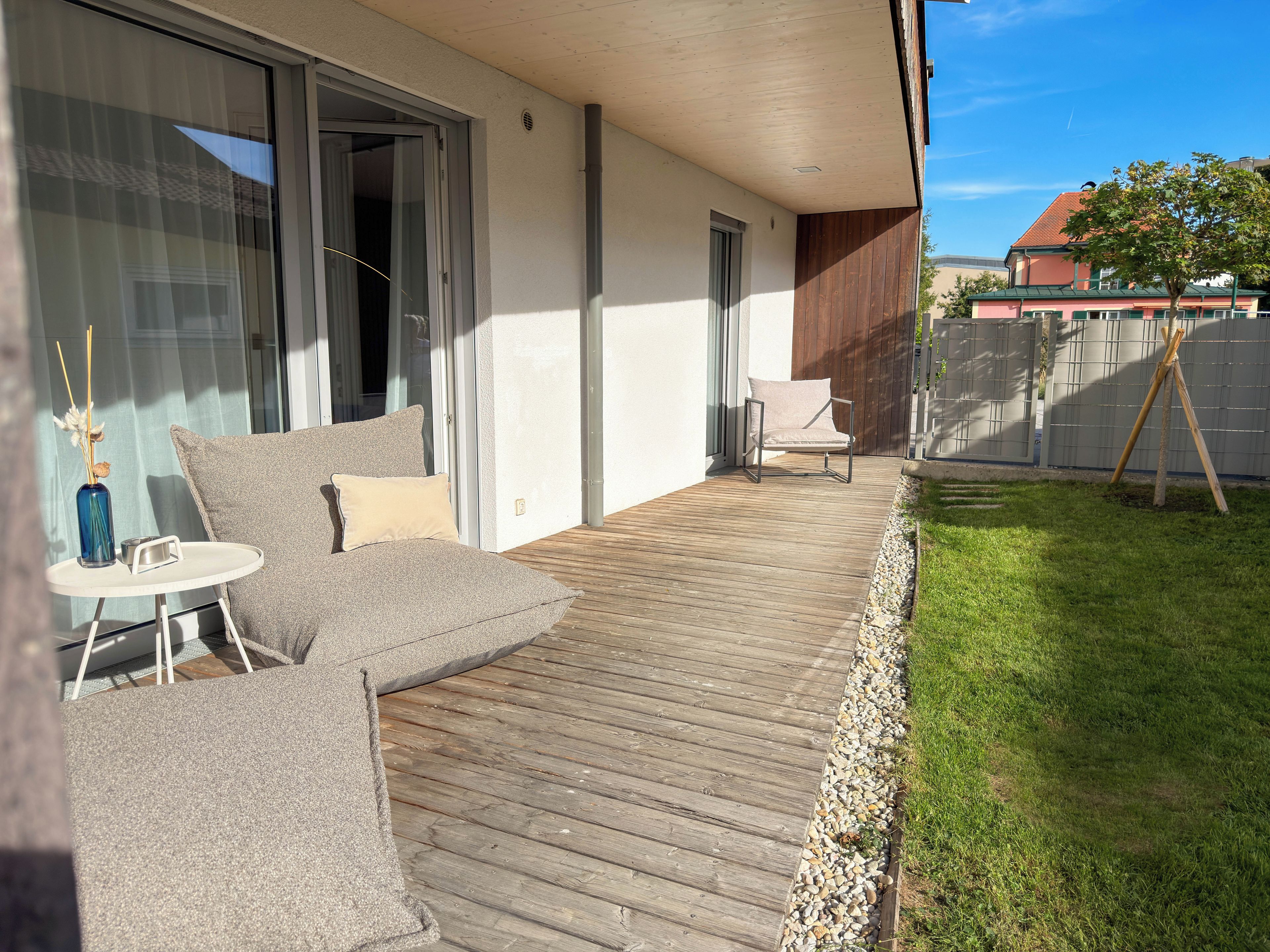 Une terrasse en bois avec un canapé et un fauteuil, une petite table avec un vase, et une zone herbeuse délimitée par une clôture.
