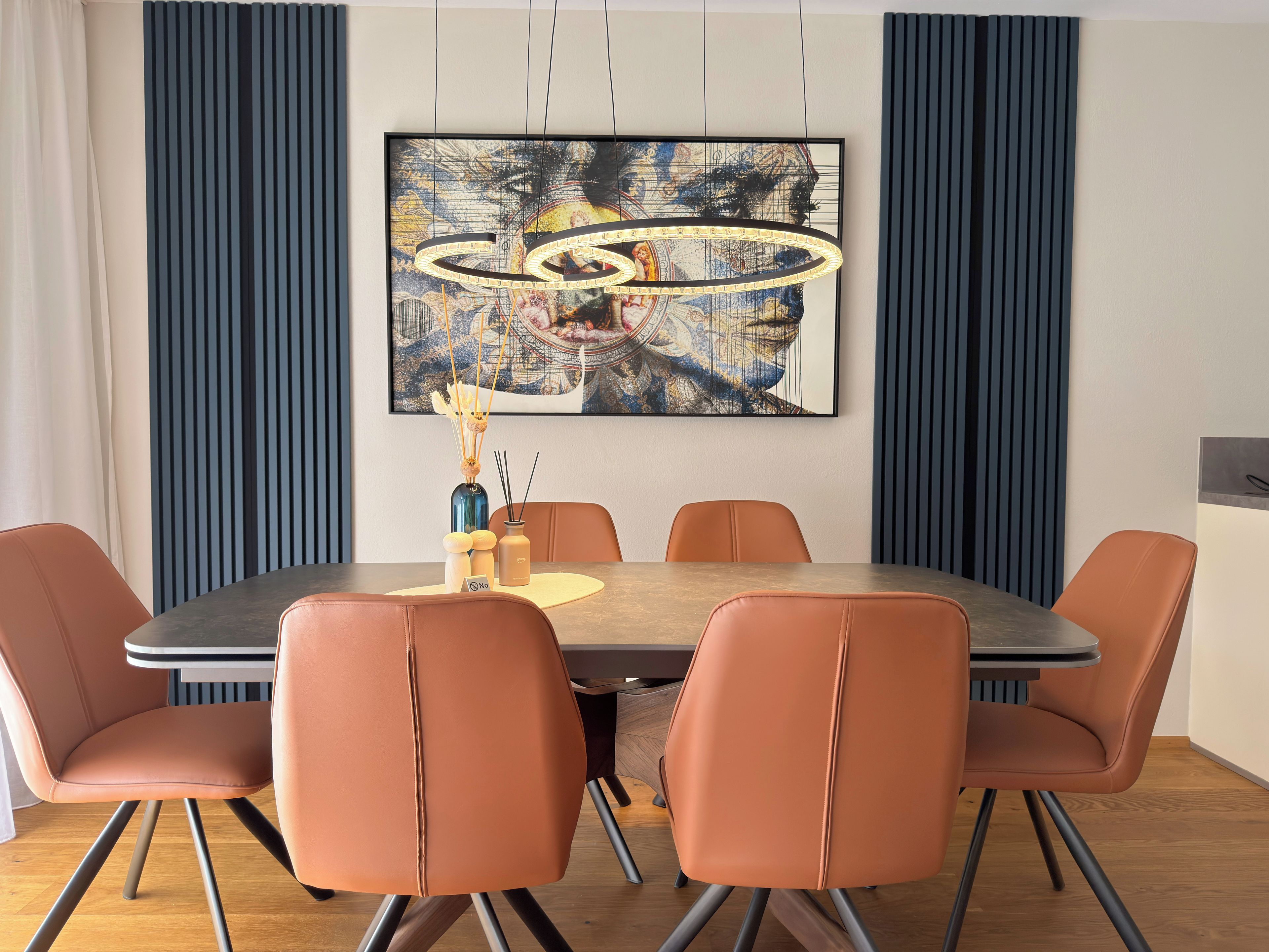 A dining area with a round table, six chairs, a painting on the wall, and a decorative vase with candles.
