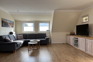 A modern living room with a sectional sofa, a small coffee table, a television on a wooden cabinet, and a window with a view. - Apartment in Cuxhaven with swimming pool (Cuxhaven)