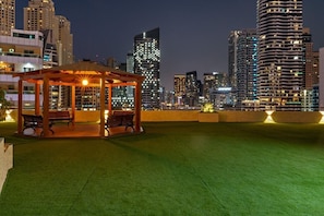 A rooftop garden with a wooden gazebo, artificial grass, and city skyline views at night. - Bright 1BR in Dubai Marina with Balcony, Pool and Gym (Dubai)