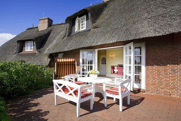 A traditional thatched-roof cottage with a brick exterior, a patio with white furniture, and a small garden area. - Holiday home in Sylt with 3 bedrooms (Sylt \/ Rantum)