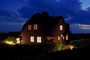 A large brick house with multiple windows, a chimney, and a well-lit entrance. - Holiday home in Sylt with 3 bedrooms (Sylt \/ Rantum)