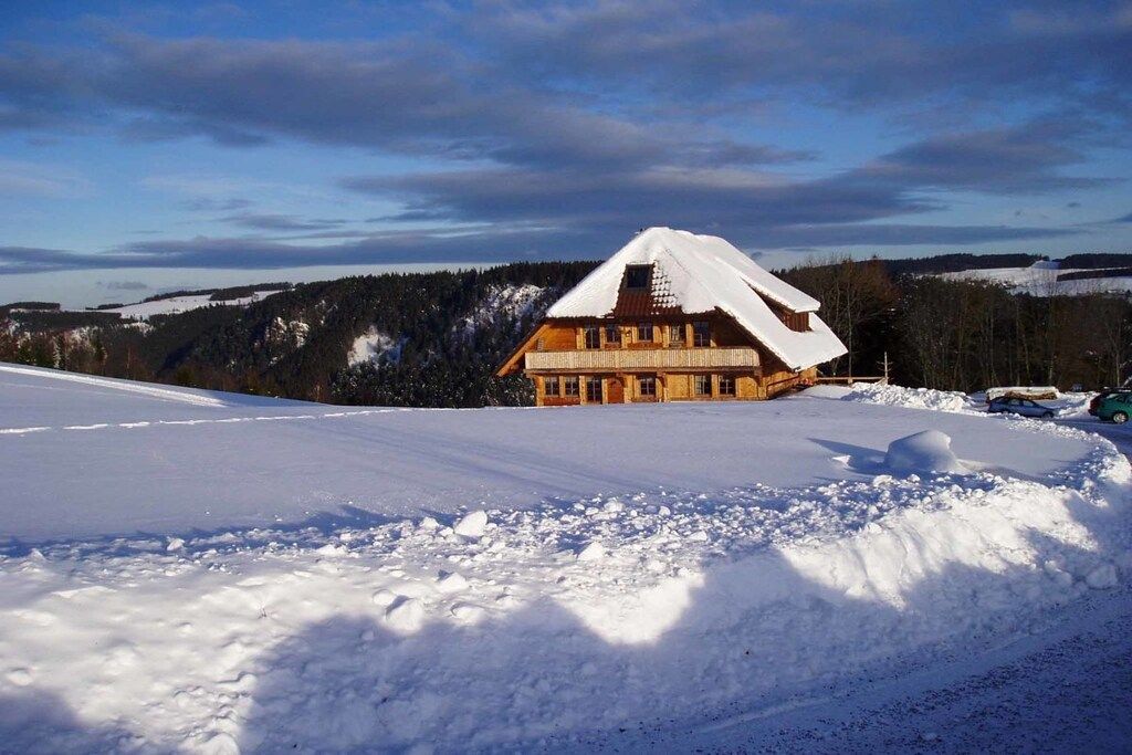 Eine Holzchalet mit verschneitem Dach, umgeben von verschneiter Landschaft und bewaldeten Hügeln.