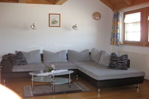 Ein Wohnzimmer mit grauem Ecksofa, einem Glastisch, einem gerahmten Bild an der Wand und einem Fenster mit Vorhängen.