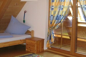 Ein kleiner, gemütlicher Raum mit schrägem Dach, einem Bett mit Kissen, einem kleinen Nachttisch aus Holz und einem Fenster mit kariertem Vorhang.
