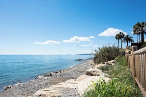 A coastal landscape with a pebble beach, a wooden fence, and palm trees. - Ocean Pearl | Sea View - Pool - Gym (Estepona)