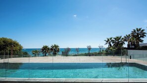 Cielo azul despejado, piscina con barrera de vidrio y vista al mar con palmeras.