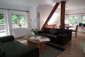Un salon avec un canapé, une table basse sur laquelle se trouvent un livre et des fleurs, un coin repas avec une table et des chaises, et une vue sur la verdure à l’extérieur.