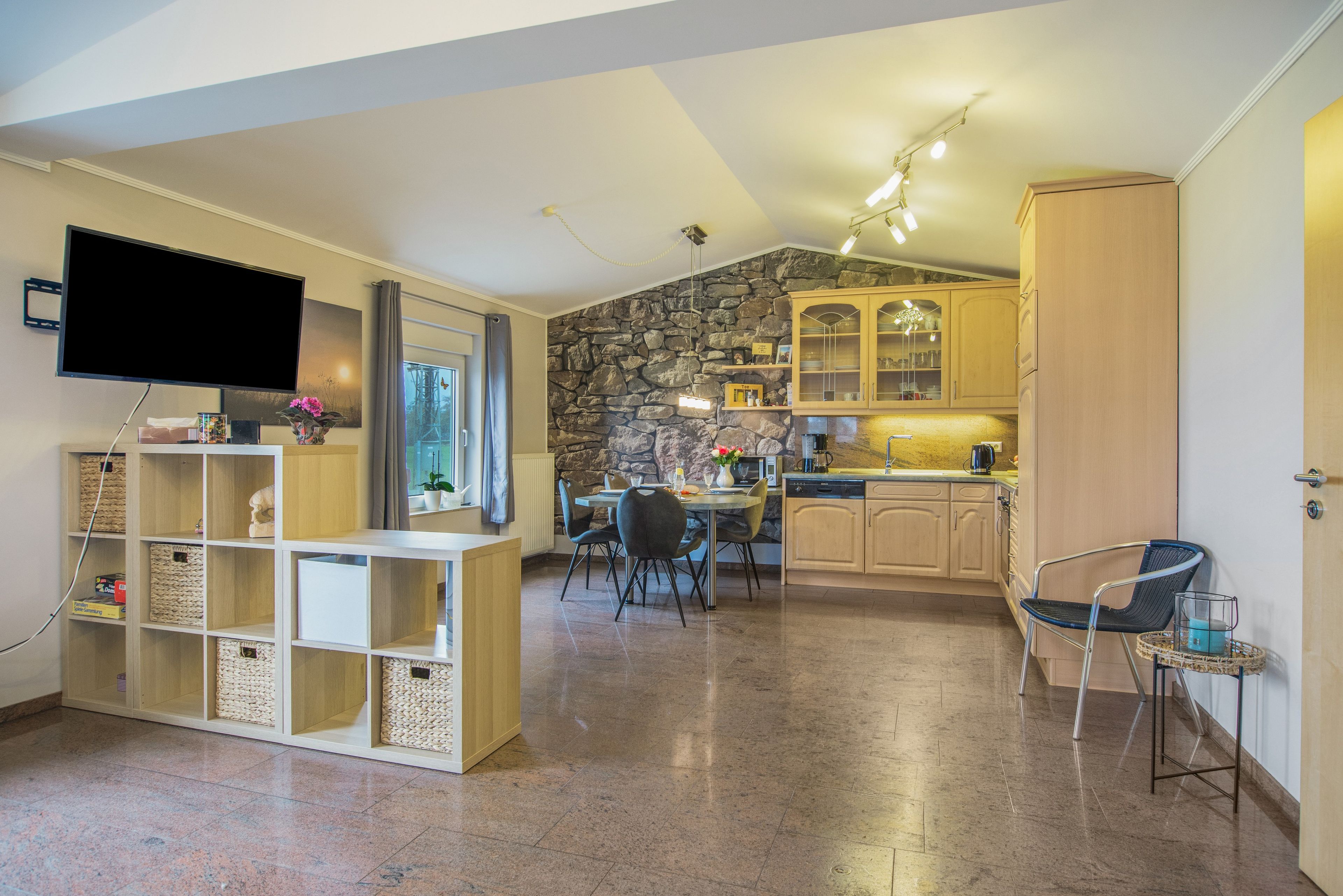 A modern kitchen with stone wall accents, a dining area with chairs, and a flat-screen TV mounted on the wall.