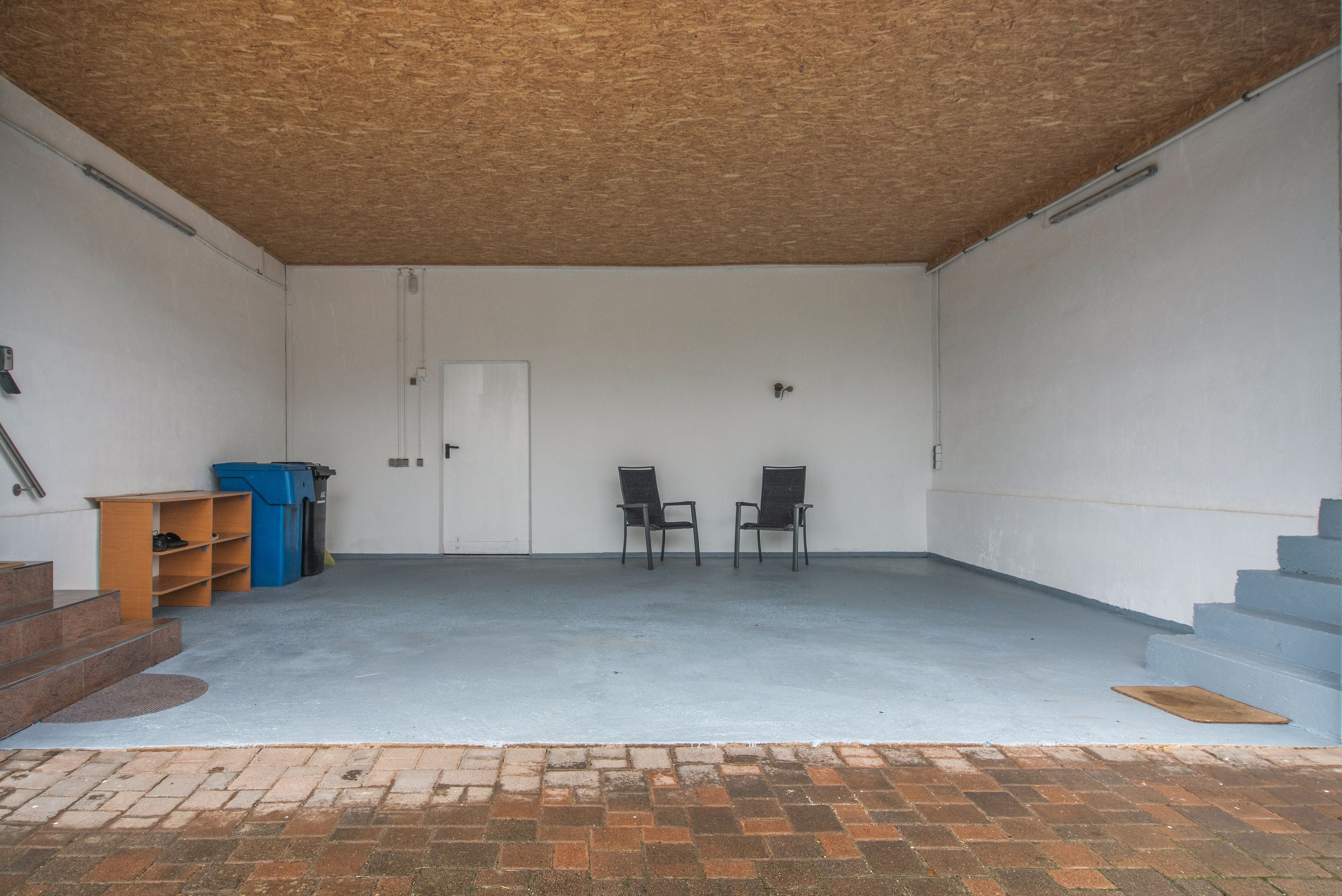 A garage with two chairs, a blue trash bin, and a wooden shelf.