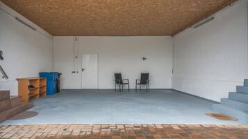 A garage with two chairs, a blue trash bin, and a wooden shelf.
