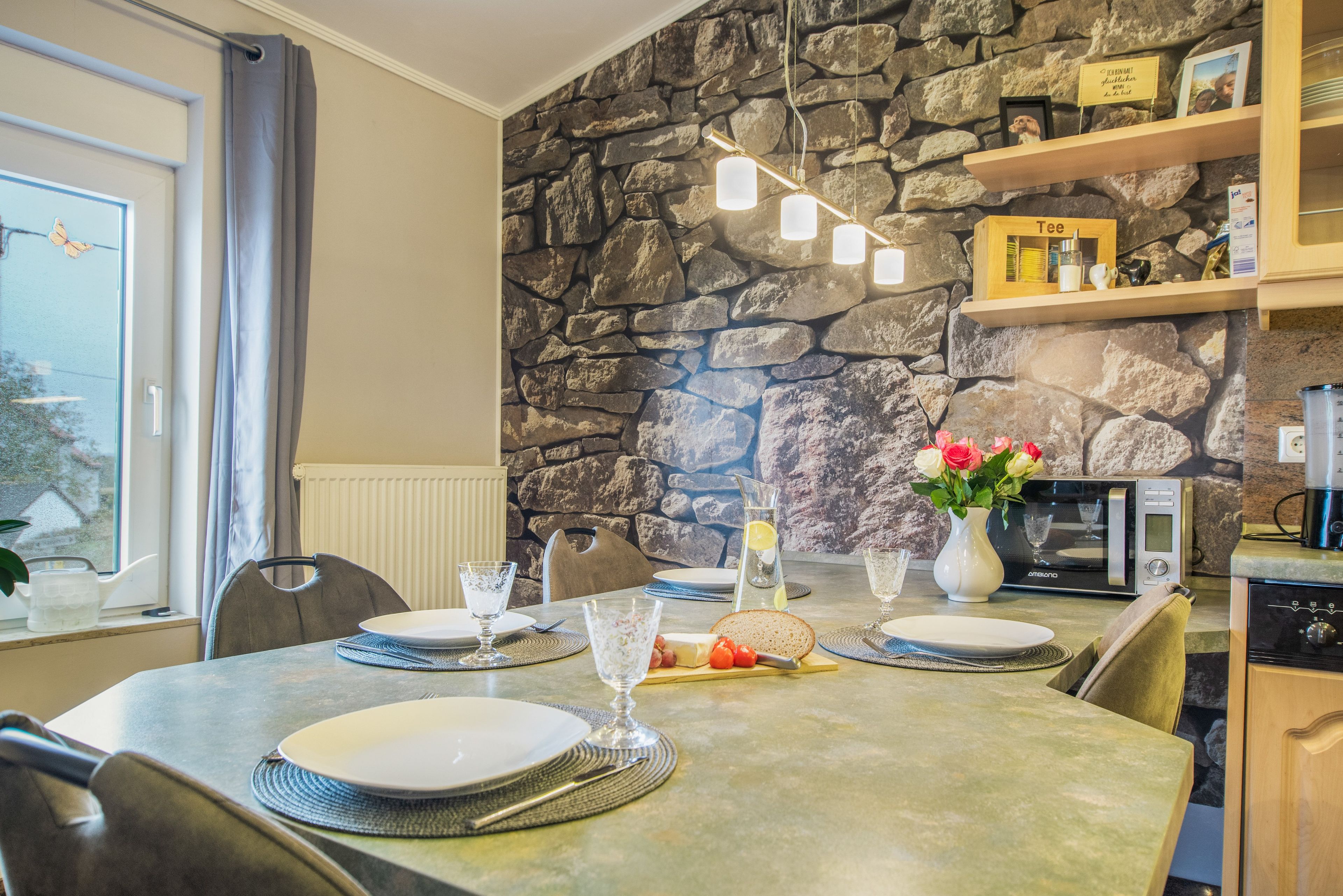 A dining area with a stone wall, a table set for four, and a kitchen area with a microwave and a vase of flowers.