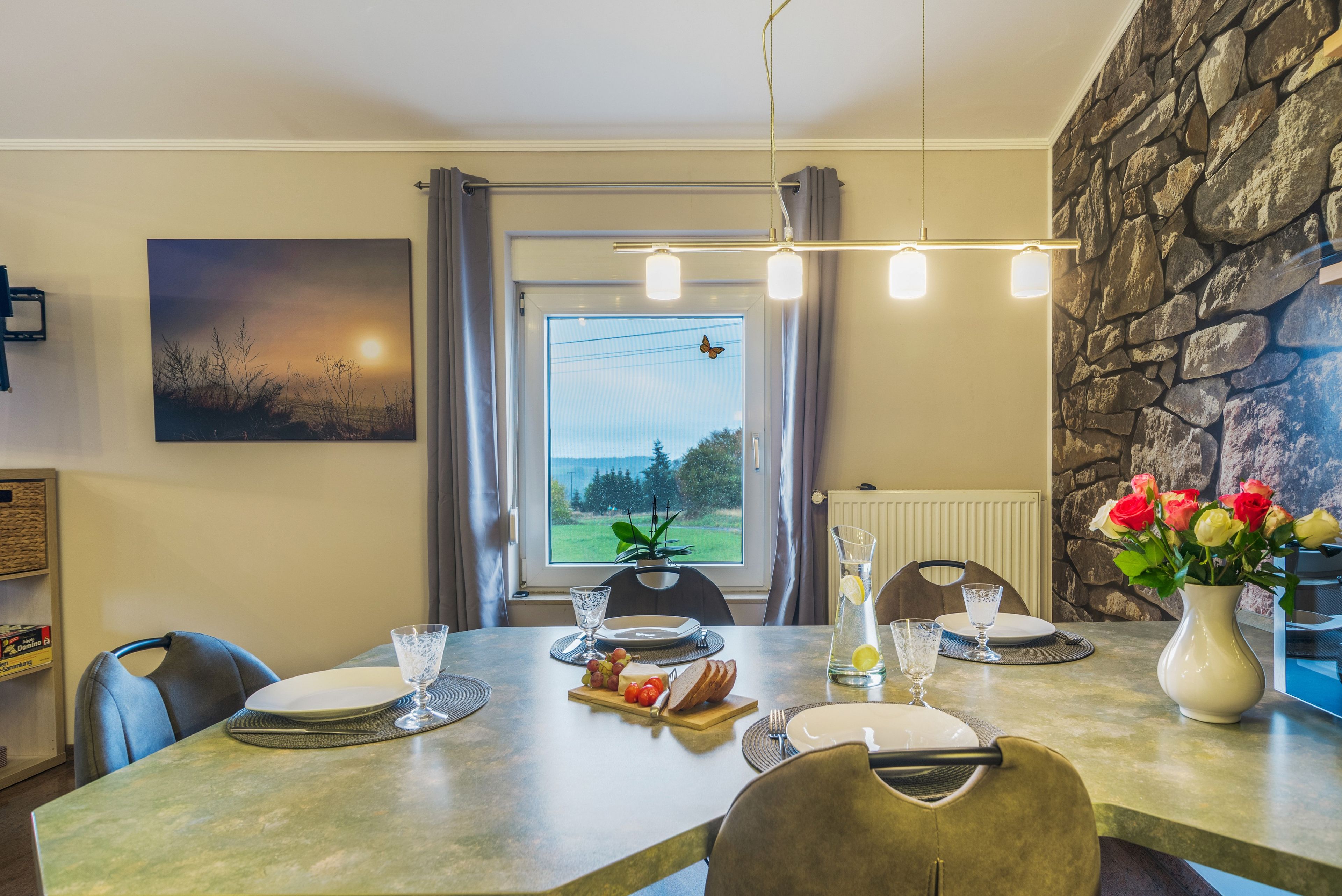 A dining area with a table set for two, a view of the outdoors through a window, and a stone wall.
