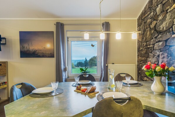 A dining area with a table set for two, a view of the outdoors through a window, and a stone wall.