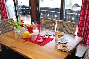 A breakfast table set with a red placemat, a glass pitcher, a glass of orange juice, a mug, a plate with eggs, a plate with a croissant, and a vase with flowers. - Holiday apartment Auszeit 3 (Obermaiselstein)