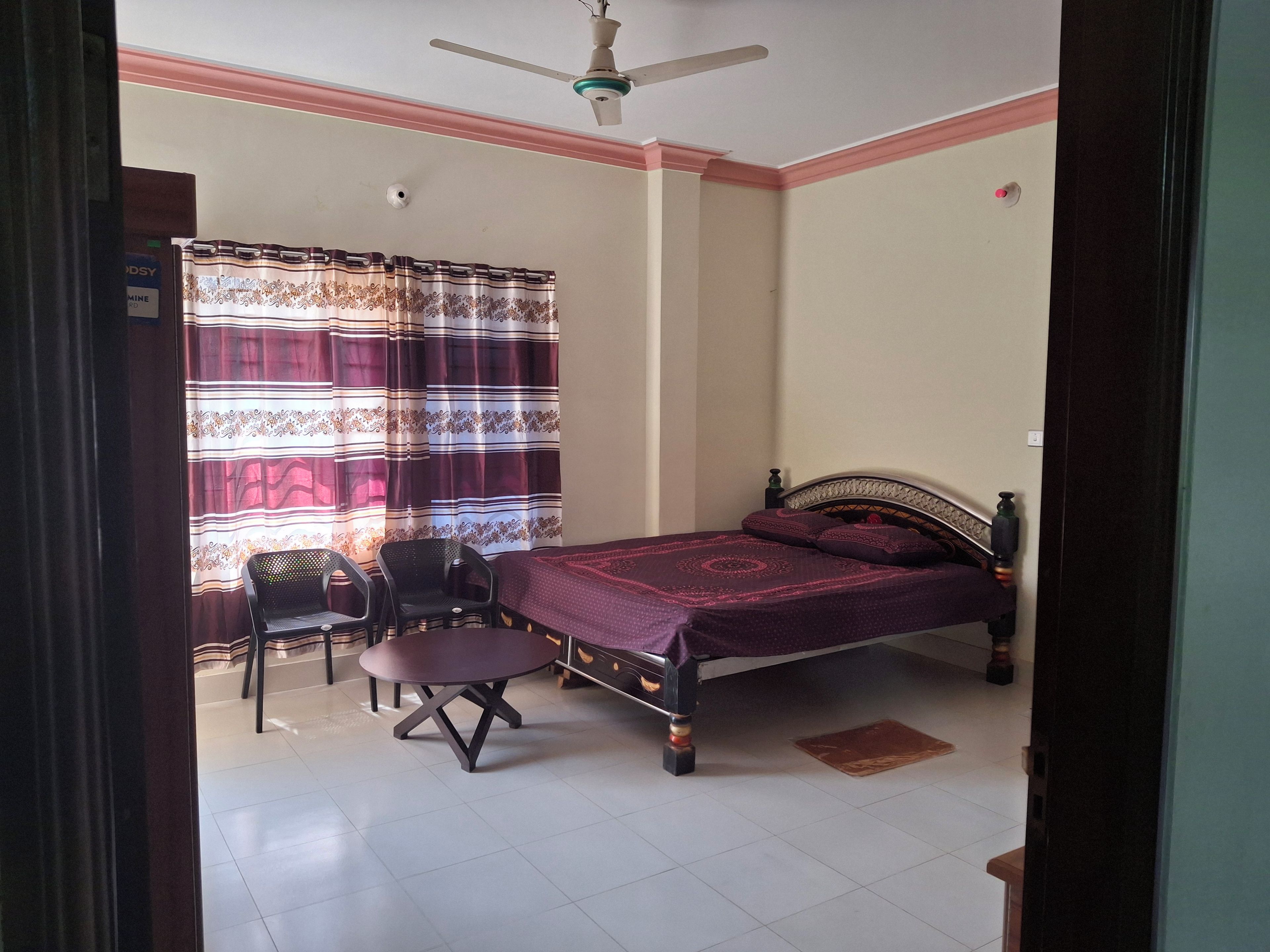 A bedroom with a bed, a chair, a table, a ceiling fan, and a window with curtains.