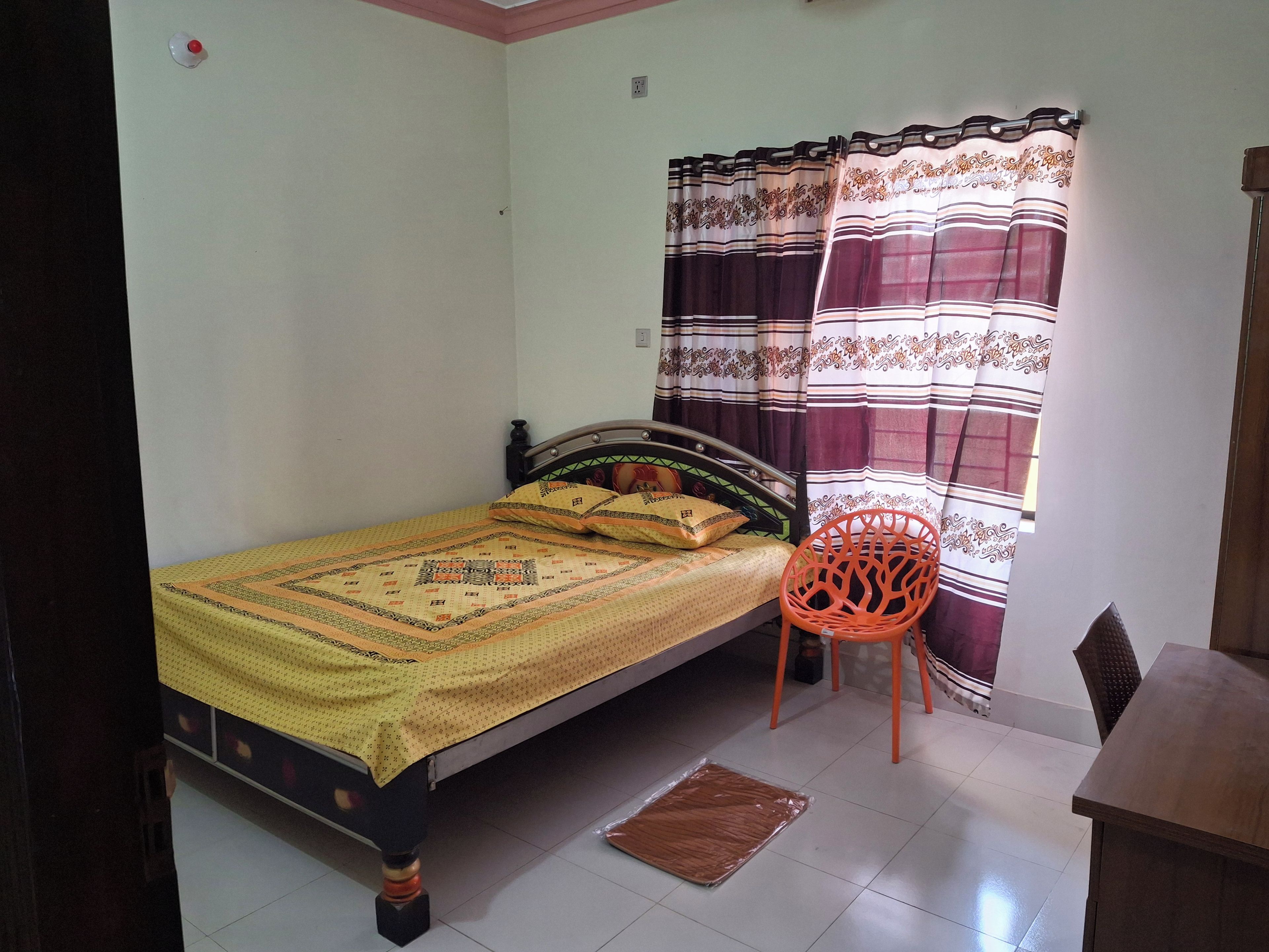 A single bed with a patterned bedspread, a chair, and a table in a room with plain walls and a tiled floor.