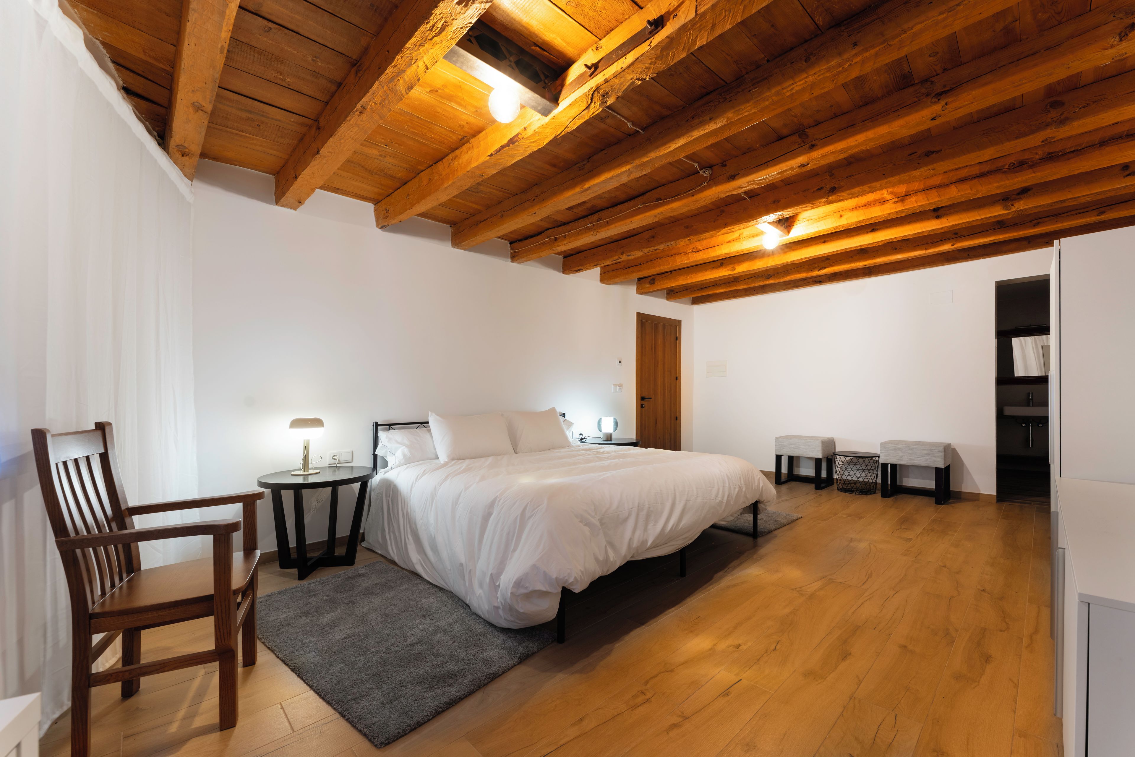 Un dormitorio con techo de madera, una cama, una silla, una mesita de noche y una pequeña mesa con lámpara.