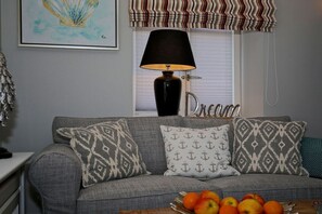 Ein Wohnzimmer mit grauem Sofa, gemusterten Kissen, einer Lampe und einer Obstschale.
