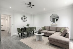 A modern living room with a sectional sofa, a dining table with chairs, a round coffee table, a wall clock, and a ceiling light fixture. - Stylish 3-Bed | Ebbsfleet & Bluewater | Parking (Northfleet)