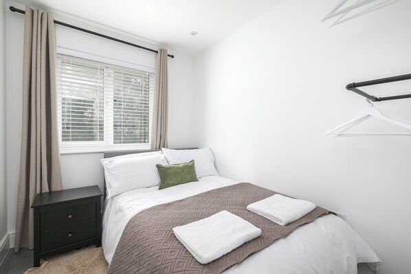 A bedroom with a bed, a nightstand, a window with curtains, and a wall-mounted hanger. - Stylish 3-Bed | Ebbsfleet & Bluewater | Parking (Northfleet)