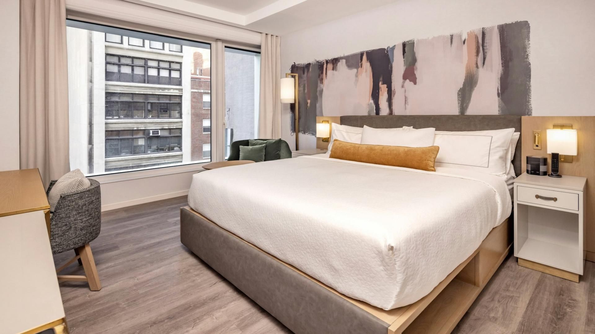A hotel room with a large bed, a desk, a chair, a nightstand, and a view of the city through the window.