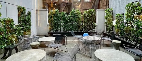 An outdoor seating area with round tables and metal chairs, surrounded by greenery and brick walls.