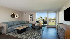 A modern living room with a grey sofa, a coffee table, a patterned rug, a chair, a TV, and a view of a golf course through large sliding doors. - Big Island Ocean Tower Stay with Partial Ocean View, Pools & Waikoloa Resort Fun (Waikoloa Village)