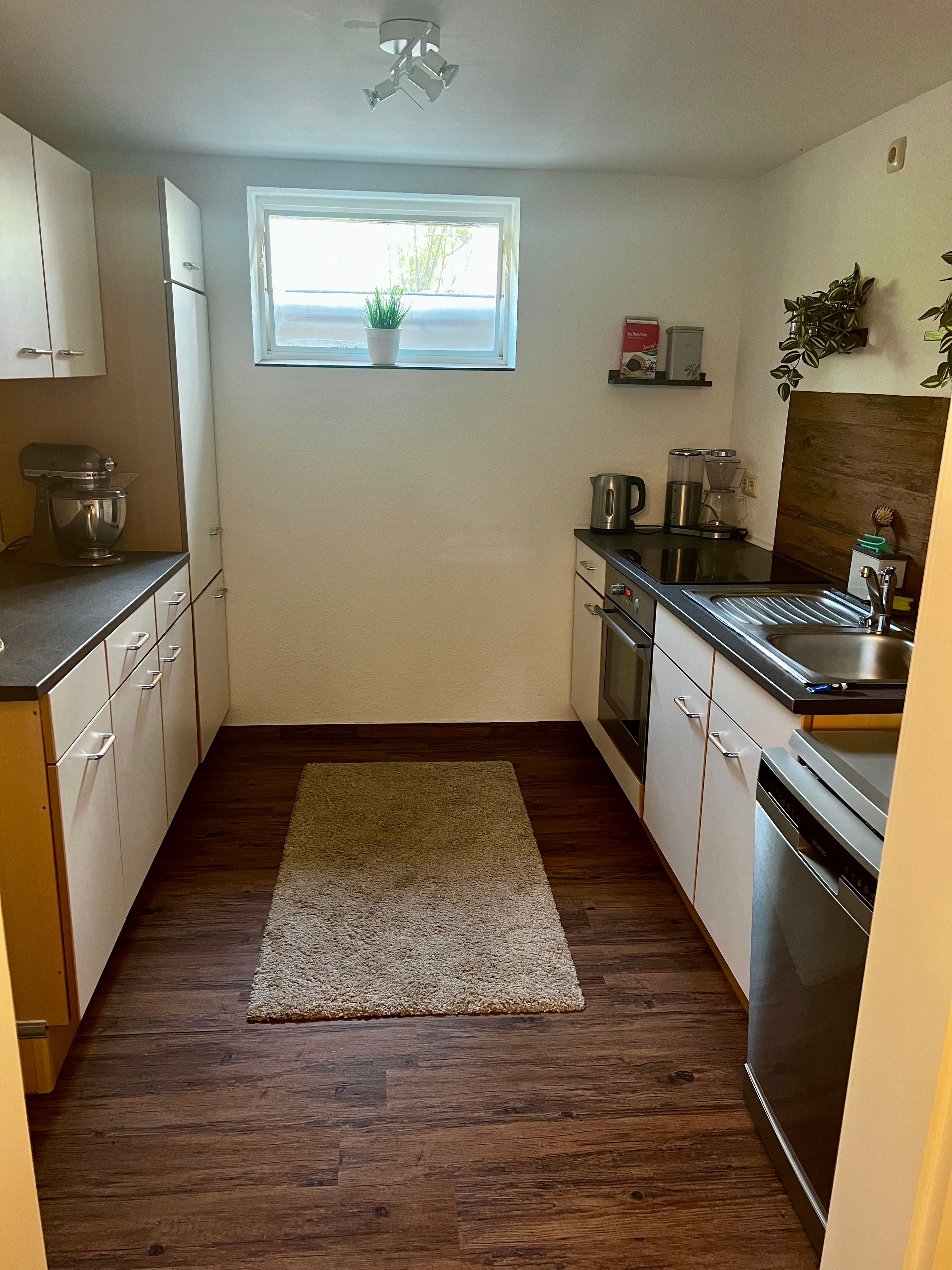 A kitchen with wooden flooring, white cabinets, a built-in oven, a dishwasher, and a window with a plant.