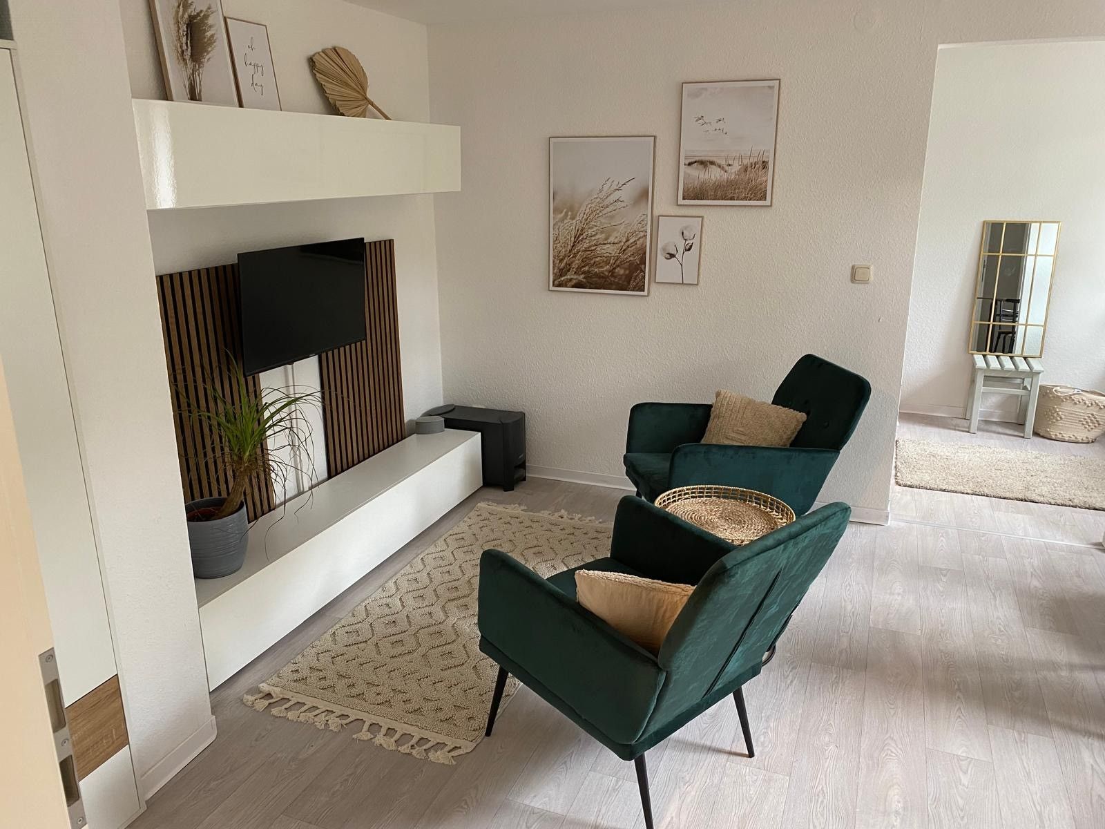 A modern living room with a flat-screen TV, two green armchairs, a patterned rug, and framed artwork on the wall.