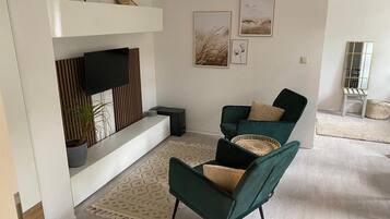 A modern living room with a flat-screen TV, two green armchairs, a patterned rug, and framed artwork on the wall.