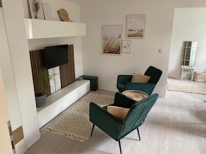 Una sala de estar moderna con un televisor de pantalla plana, dos sillones verdes, un tapete estampado y obras de arte enmarcadas en la pared.