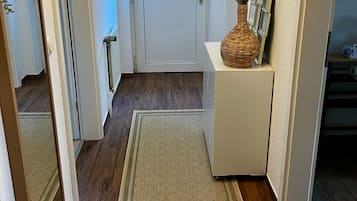 A hallway with a wooden floor, a white door with a glass panel, a white console table with a woven basket, and a mirror on the left.