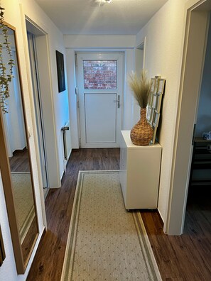 A hallway with a wooden floor, a white door with a glass panel, a white console table with a woven basket, and a mirror on the left.