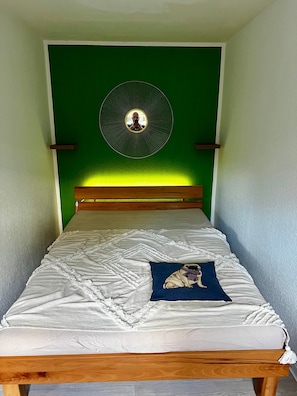 Una cama con colcha blanca, almohada azul con imagen de perro y un adorno circular en la pared.