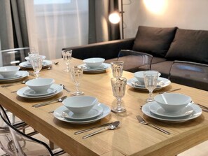 Una mesa de comedor dispuesta con platos blancos, fuentes y cristalería transparente, con un sofá moderno y una lámpara al fondo.