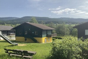 A building with a green and yellow exterior, a slide, and a picnic table in a grassy area with hills in the background. - by the lake with lake and Bavarian Forest views (upper floor) (Arrach\/ Kummersdorf)