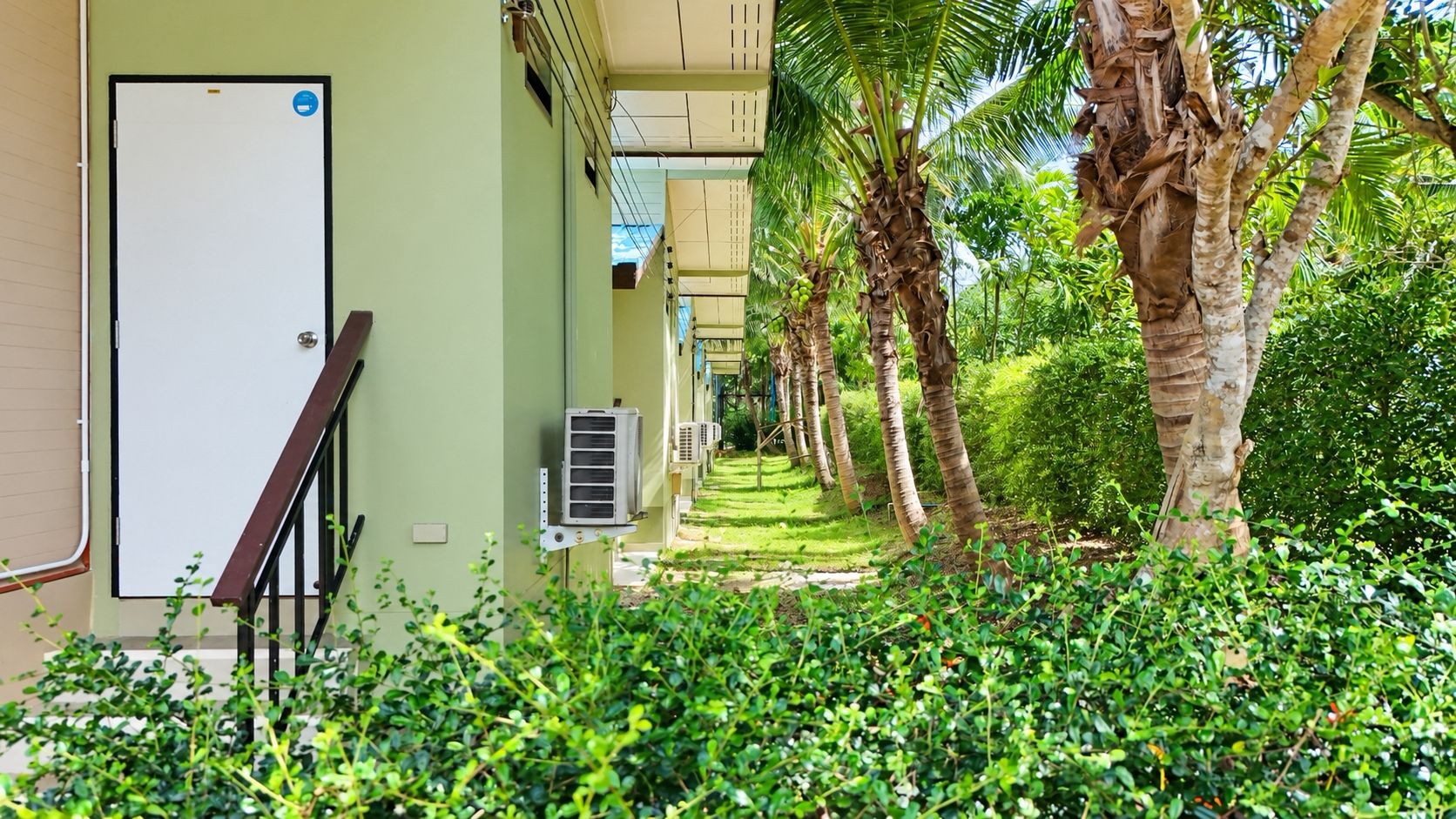 A corridor with a door, air conditioning units, and a pathway lined with palm trees and greenery.