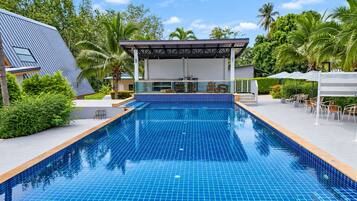 Una piscina con plataforma de baldosas, rodeada de follaje y una zona de estar cubierta.