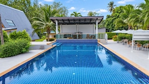 A swimming pool with a tiled deck, surrounded by greenery and a covered seating area.