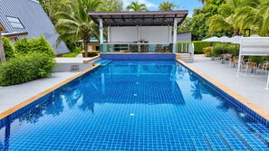 A rectangular swimming pool with blue tiles, surrounded by a covered seating area with chairs and tables, and a building with a sloped roof in the background. - Flow Krabi resort (Mueang)