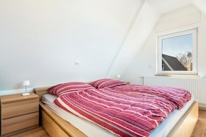 A bedroom with a sloped ceiling, a window, a bed with striped bedding, a bedside table with a lamp, and a radiator. - Freddy's Beach House (Cuxhaven)