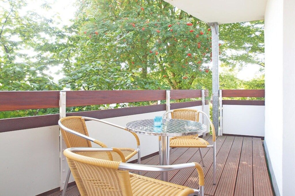 A balcony with wicker chairs and a glass table, surrounded by a wooden deck and a white wall.