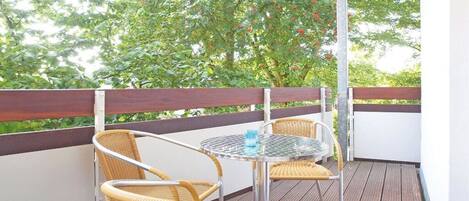 A balcony with wicker chairs and a glass table, surrounded by a wooden deck and a white wall.