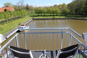 A view from a balcony overlooking a pond with a boat, surrounded by greenery and a fence. - At the canal footbridge (Krummhörn-Greetsiel)